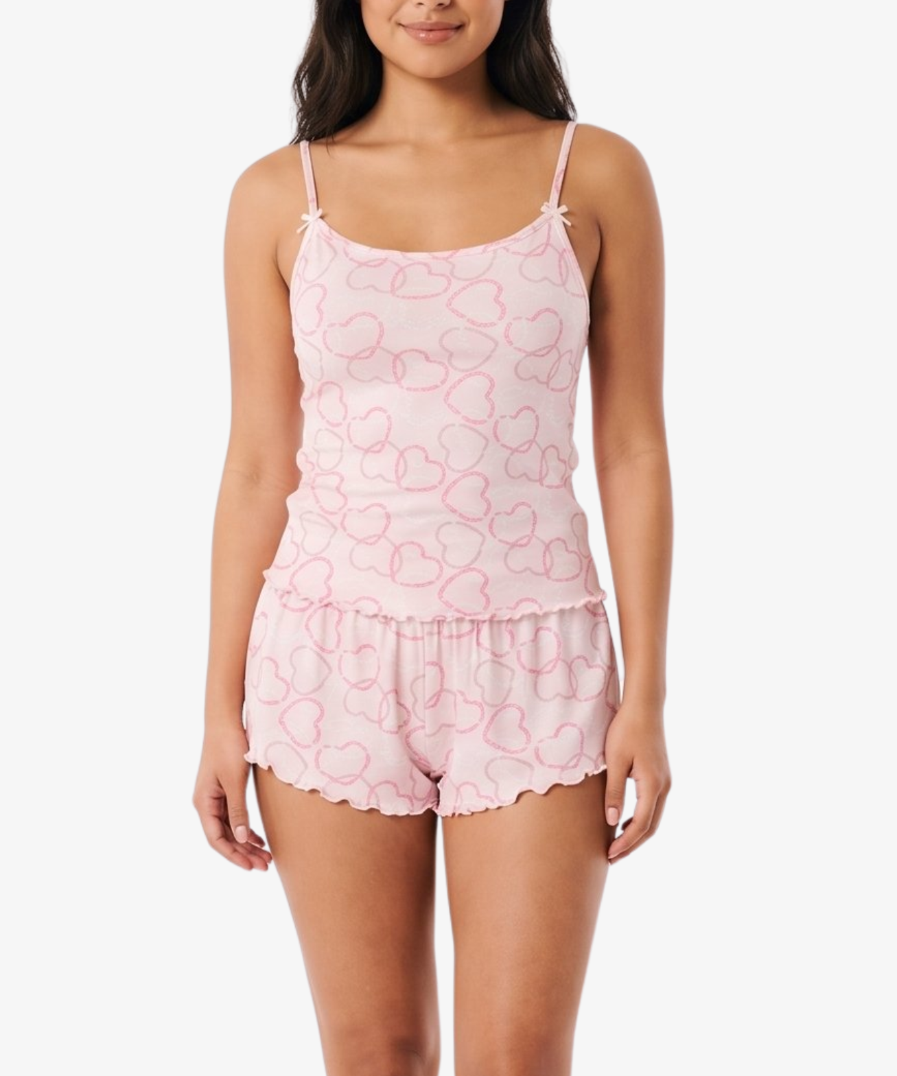 A woman modeling a light pink two-piece pajama set with a subtle heart pattern, consisting of a spaghetti-strap camisole top and matching ruffled shorts, against a plain white background.