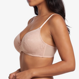A woman modeling a light beige/nude lace bra and matching lace underwear against a plain light gray studio background.