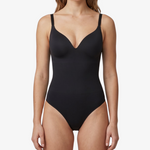 A woman modeling a black one-piece shapewear with thin shoulder straps and a deep V-neckline, photographed against a clean light gray studio background.