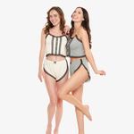 Two women in matching pajama sets behind a white background. One woman is wearing a cream pajama set and the other woman is wearing a gray pajama set.